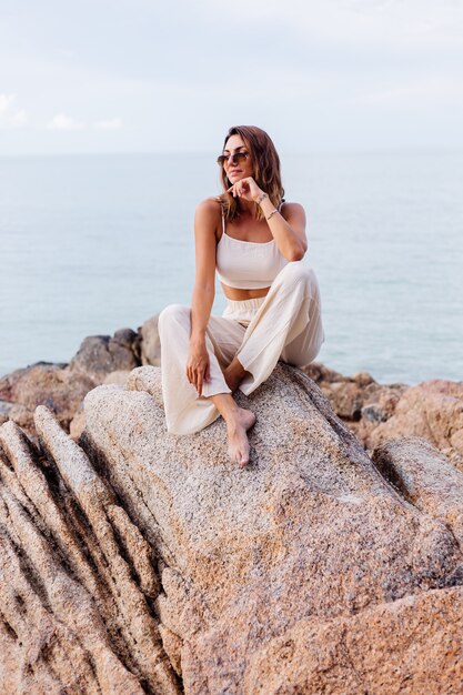 portrait of young calm happy caucasian fit slim woman in crop cami top and pants set sits alone on rocky tropical beach at sunset