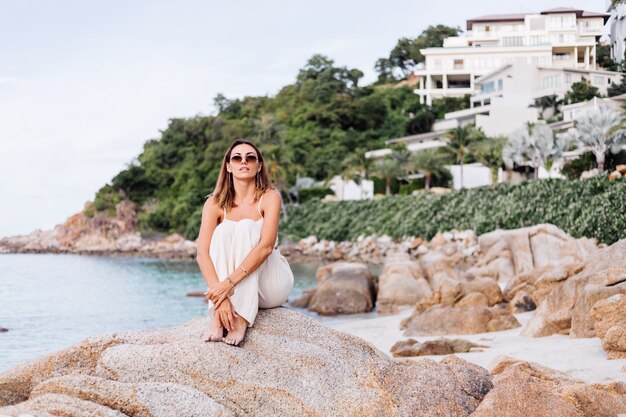 portrait of young calm happy caucasian fit slim woman in crop cami top and pants set sits alone on rocky tropical beach at sunset