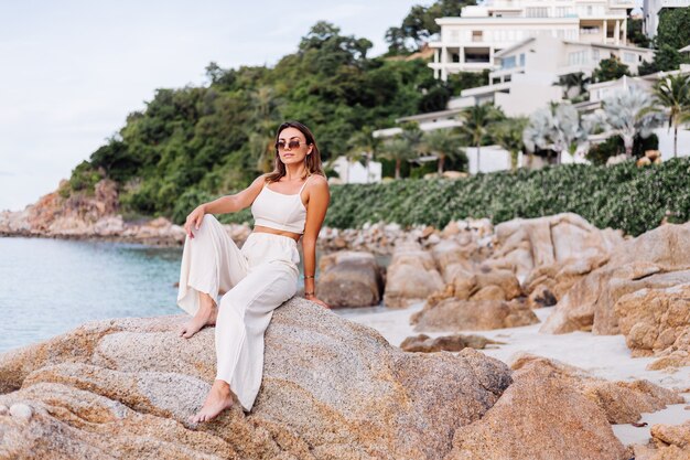 portrait of young calm happy caucasian fit slim woman in crop cami top and pants set sits alone on rocky tropical beach at sunset