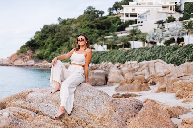 portrait of young calm happy caucasian fit slim woman in crop cami top and pants set sits alone on rocky tropical beach at sunset