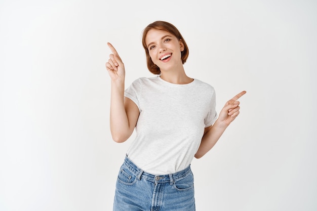 Portrait of young carefree woman pointing at two sides directions. Girl showing left and right ways, standing on white wall and smiling