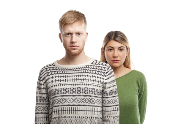 Portrait of young Caucasian couple wearing casual clothes posing, having serious facial expressions: blonde woman standing at white wall behind her unshaven boyfriend dressed in sweater