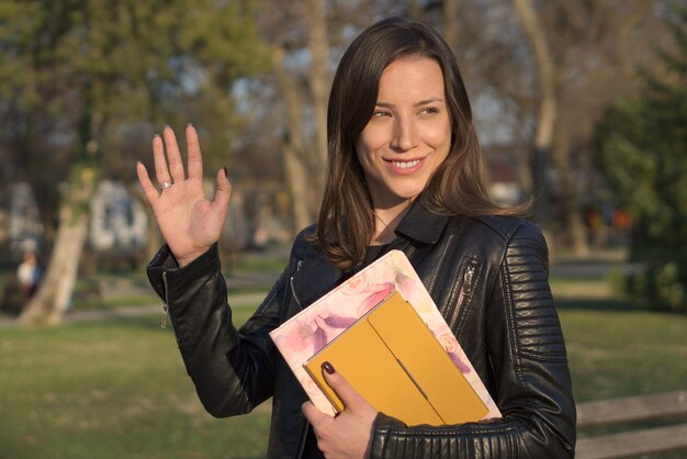 Portrait of young caucasian female wearing leather jacket holding notebooks smiling and waving