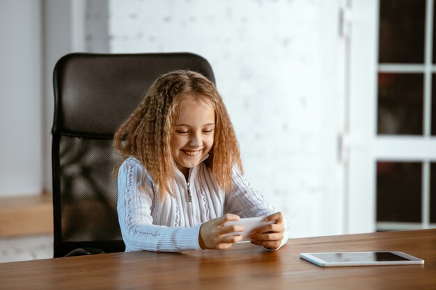 Portrait of young caucasian girl looks dreamful, cute and happy. Looking up, sitting indoors at the wooden table with tablet and smartphone. Concept of future, target, dream to buy, visualisation.