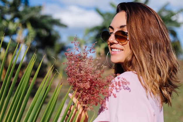 Portrait of young caucasian tanned woman in romantic pink dress round earings silver bracelet and sunglasses with wild flowers