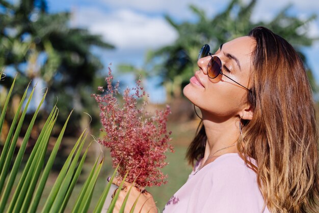 Portrait of young caucasian tanned woman in romantic pink dress round earings silver bracelet and sunglasses with wild flowers