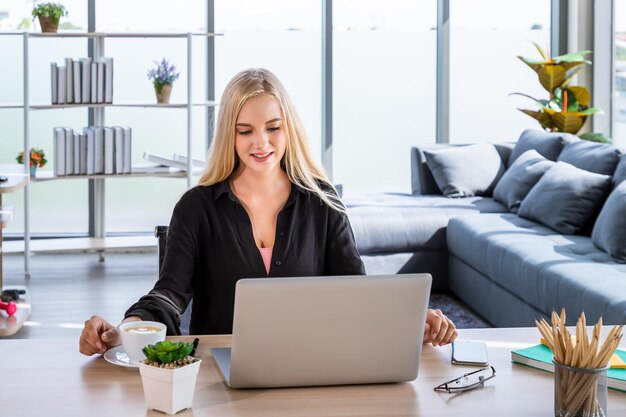 Portrait of young Caucasian woman with blonde hair freelancer working at home sitting at desk in living room using typing laptop computer