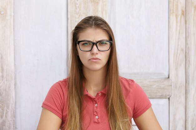 Portrait of young Caucasian woman with long loose hair wearing stylish glasses and polo shirt having upset and unhappy look, frowning and making wry face, sitting against wooden wall