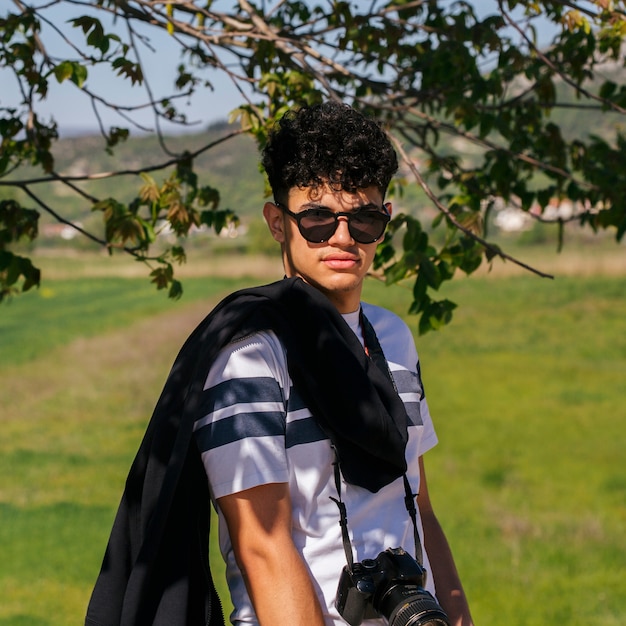 Портрет молодого очаровательного человека носить солнцезащитные очки и цифровой фотоаппарат, глядя на камеру