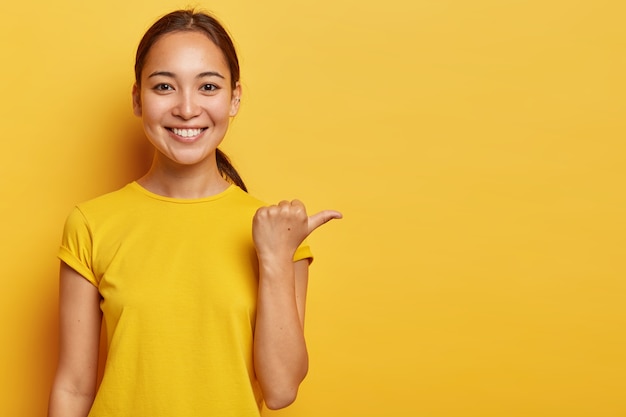 Portrait of young cheerful asian female points away with thumb, happy face expression, demonstrates copy space for advertisement, has pleasant appearance, wears bright yellow clothes.