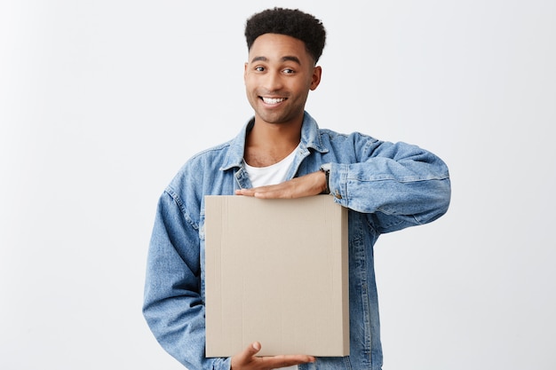 Portrait of young cheerful attractive dark-skinned man with afro hairstyle in white shirt and blue jacket holding paper box in hands with bright smile and happy expression