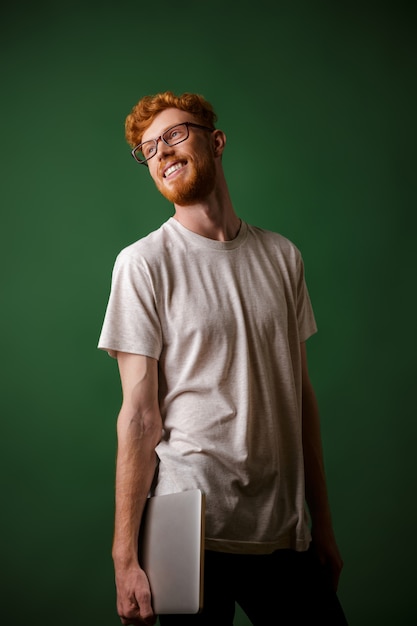 Portrait of young cheerful bearded readhead hipster, holding laptop, looking away