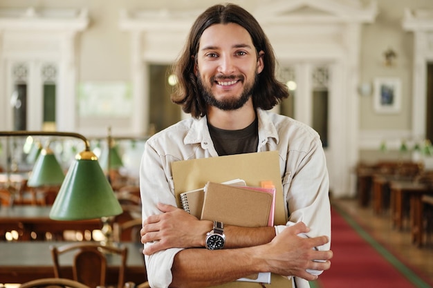 Portrait of young cheerful casual male student with notepads joyfully looking in camera in library of university