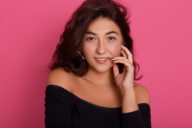 Portrait of young cheerful fresh woman with flirting smile, posing isolated over pink wall with finger on lips, dark haired female .