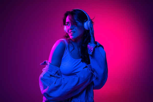 Portrait of young cheerful girl listening to music in headphones isolated over gradient pink background in blue neon light