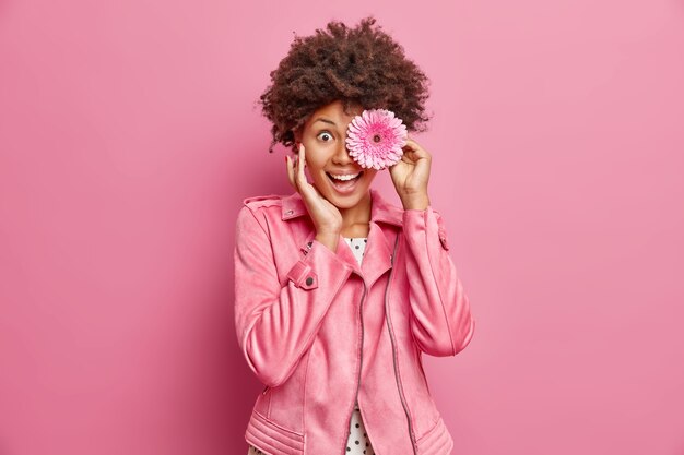 Portrait of young cheerful woman keeps pink gerbera flower in front of eye touches face gently feels very happy wears stylish jacket poses against rosy wall