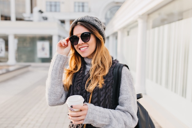 Портрет молодой городской модной женщины в современных солнцезащитных очках, теплом шерстяном свитере, вязаной шапке, улыбаясь на улице. Веселое настроение, положительные эмоции, прогулки с кофе на вынос.