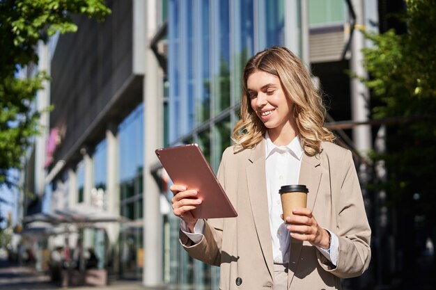 Portrait of young confident businesswoman on street drinks her coffee and looks at tablet works on h