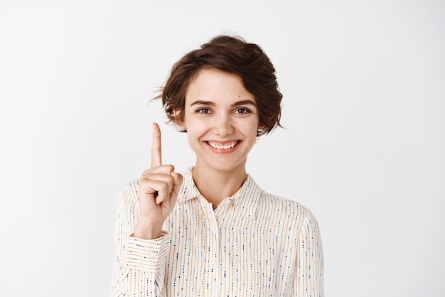 Portrait of young confident woman pointing finger up and smiling showing promo logo standing over white background in blouse