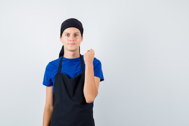 Портрет молодого повара, поднимающего сжатый кулак в футболке, фартуке и серьезного вида спереди