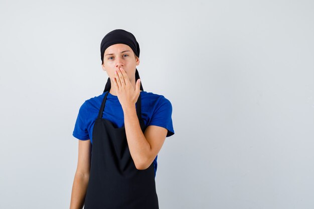 Portrait of young cook man with hand on mouth in t-shirt, apron and looking puzzled front view