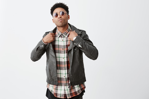 Free photo portrait of young cool dark-skinned man with afro hairdo in stylish leather jacket and glasses gesticulating with hands with mean face expression.