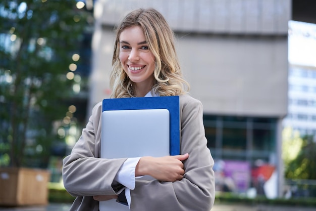 Free photo portrait of young corporate woman office manager walking on street with laptop and work folder smili