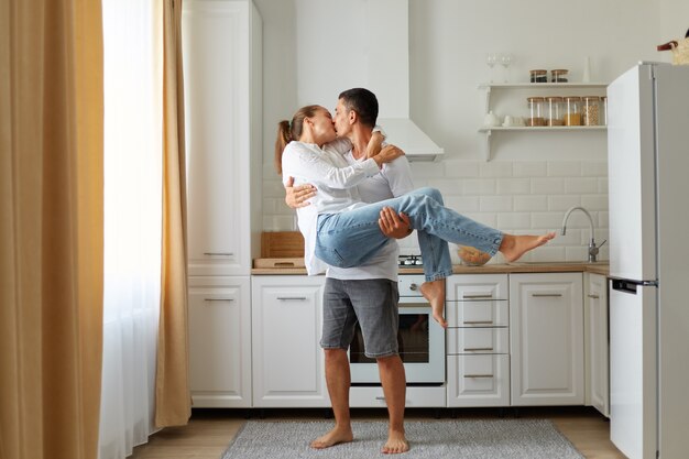 Portrait of young couple in a cosy kitchen, guy and the girl kiss and hug, man holds a woman in his arms, spending time together, honey moon, romantic feelings.