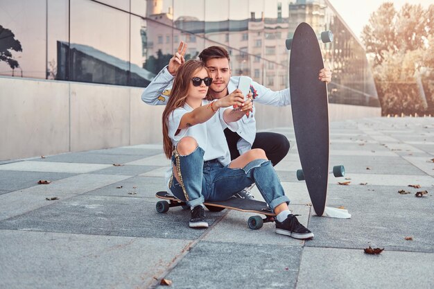 トレンディな服を着て楽しんでいる若いカップルの肖像画は、屋外で一緒に自分撮りをします。