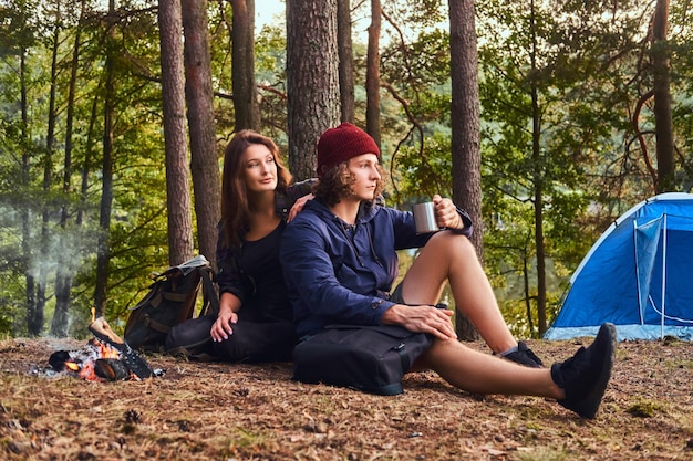 젊은 부부의 초상화 - 잘생긴 곱슬머리 남자와 숲에 함께 앉아 있는 매력적인 여자. 여행, 관광 및 하이킹 개념입니다.