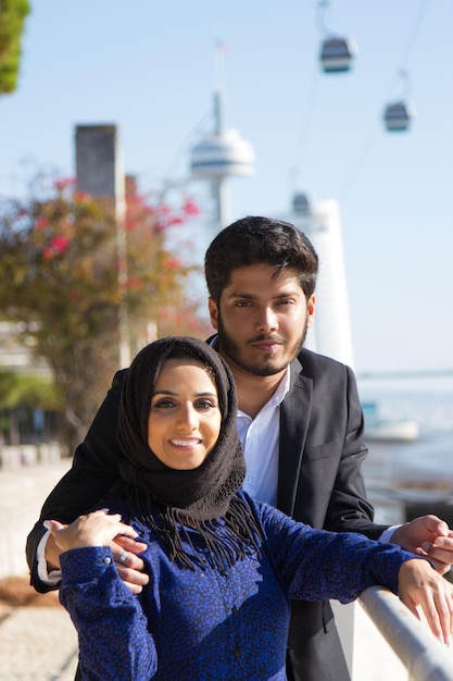 街で楽しい時間を過ごしている若いカップルの肖像画。黒のジャケットを着たイスラム教徒の男性と黒のスカーフの笑顔の女性が通りで手に手で柵の近くに立っています。ロマンス、デート、休暇のコンセプト