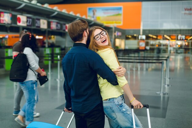 Portrait of young couple hugging in airport. She has long hair, yellow sweater, jeans and smiling to the camera. He has black shirt, pants and suitcase near. View from back.