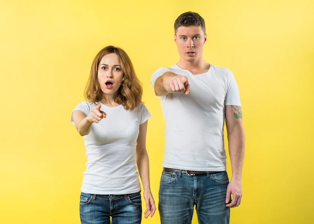 Free photo portrait of a young couple pointing fingers toward camera against yellow backdrop