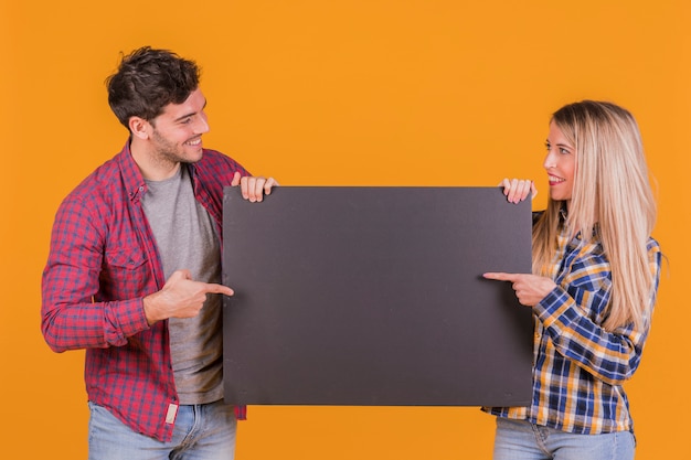 Портрет молодой пары, указывая пальцем на пустой черный плакат на оранжевом фоне