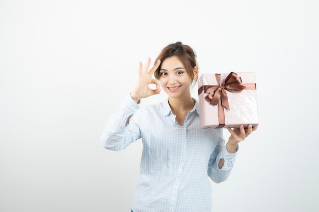 Portrait of a young cute girl doing ok gesture and holding present box with ribbon . High quality photo
