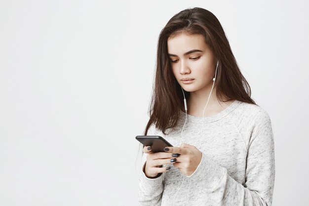 Portrait of young cute woman, looking concentrated at her phone screen while listening to music