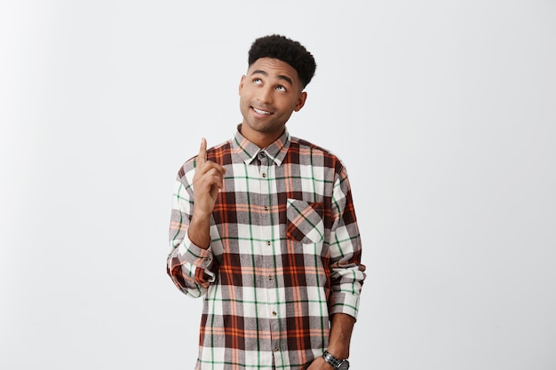 Portrait of young dark-skinned african attractive man with curly hairstyle in casual checkered shirt pointing upside with happy and relaxed expression. Copy space