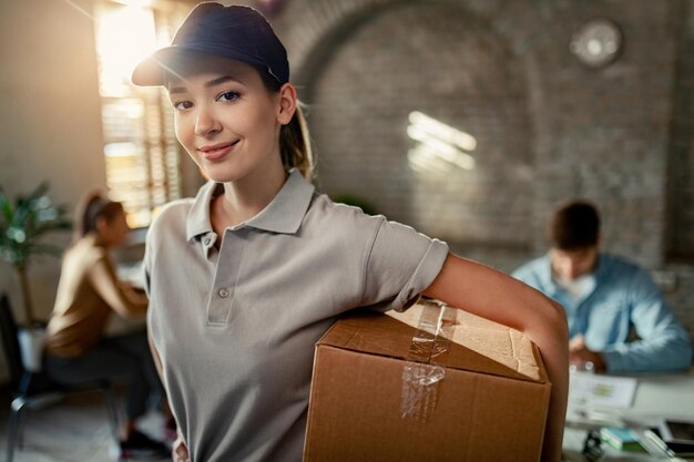 Портрет молодой доставщицы с картонной коробкой во время доставки в офис На заднем плане люди