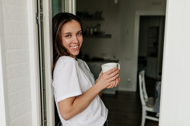 Portrait of young european woman with dark hair and healthy skin smiling with morning coffee in the modern light kitchen.