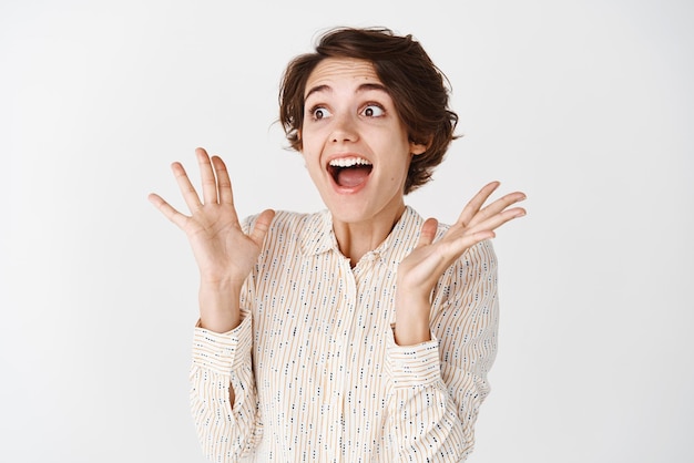 Portrait of young excited woman scream from joy and happiness looking left at empty space and gasping fascinated standing on white background