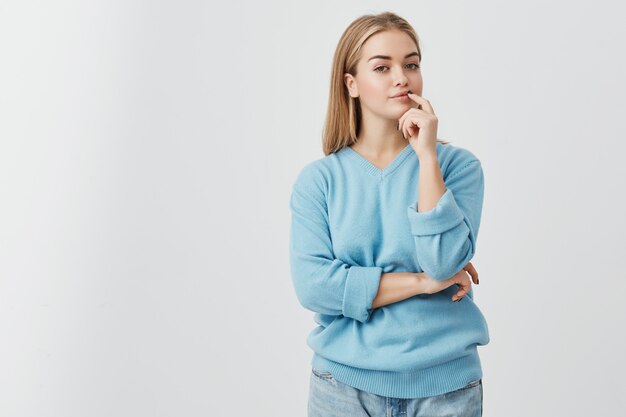 Portrait of young fair-haired european girl with healthy skin wearing blue sweater and jeans looking  with calm and thoughtful expression, thinking over about preposition she was given.