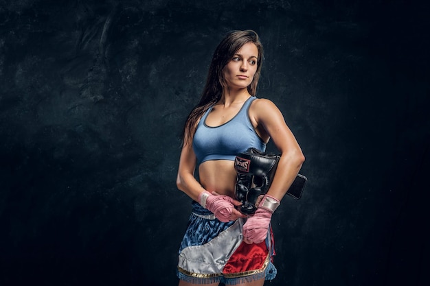 Portrait of young female boxer with her helmet in hands at dark photo studio.