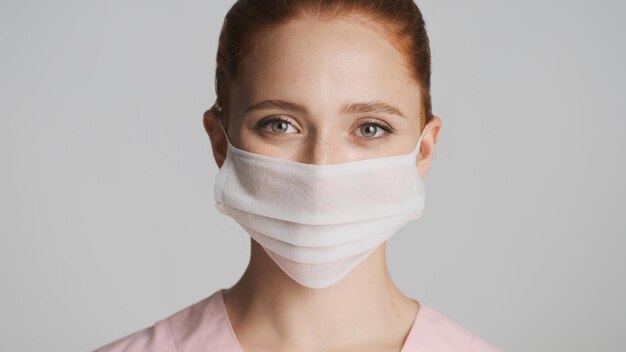 Portrait of young female doctor wearing medical mask happily looking in camera over white background Safety concept