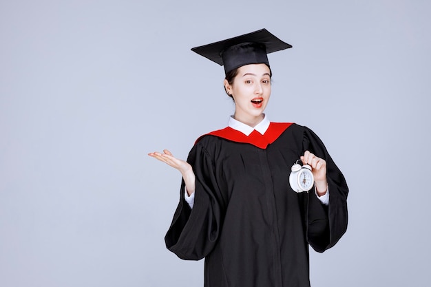 Portrait of young female graduate student holding clock to show time. High quality photo