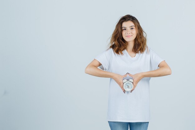 Portrait of young female holding alarm clock in white t-shirt, jeans and looking cheerful front view