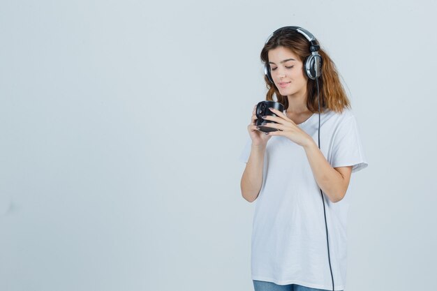 Portrait of young female holding cup of drink in white t-shirt and looking delighted front view