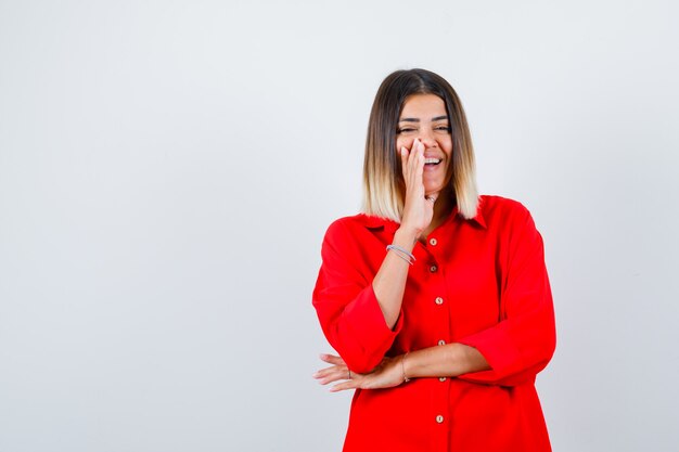 Portrait of young female holding hand on cheek in red oversized shirt and looking cheerful front view