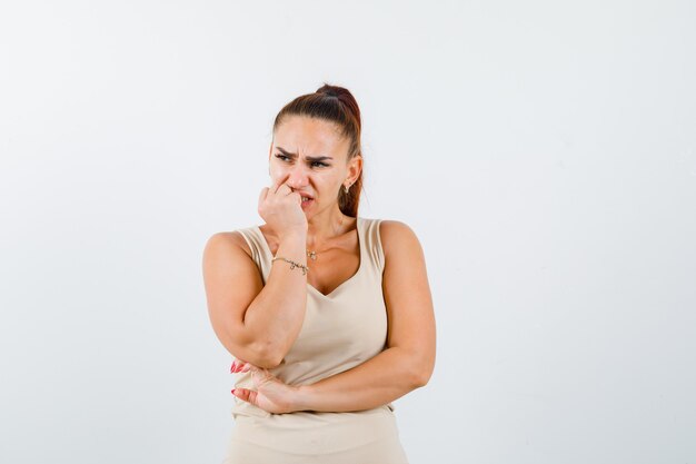 Portrait of young female holding hand on chin in beige tank top and looking scared front view