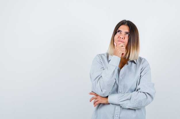 Portrait of young female holding hand on chin in oversized shirt and looking thoughtful front view