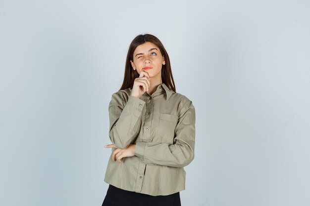 Portrait of young female holding hand on chin in shirt, skirt and looking thoughtful front view